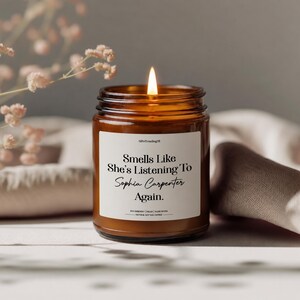 May include: An amber glass candle with a lit wick and a white label. The label says "Smells Like She's Listening To Sophia Carpenter Again." The candle sits on a neutral background with flowers.