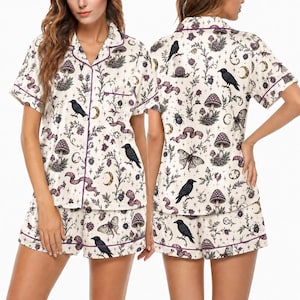 May include: A short-sleeved button-down pajama shirt and matching shorts set in an off-white fabric featuring a dark purple piping trim. The print displays black ravens, various mushrooms, moths, snails, crescent moons, and botanical foliage.