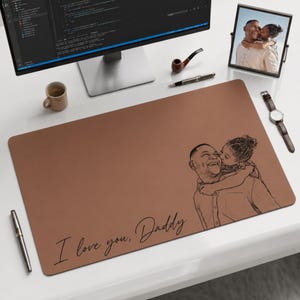 May include: A brown desk mat with a sketch of a father and child, and the text "I love you, Daddy." A computer monitor, coffee mug, pen, pipe, picture frame, and watch are also on the white desk.