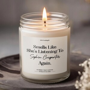 May include: A lit candle in a clear glass jar with a white label. The label says "Smells Like She's Listening To Sophia Carpenter Again." The candle is made with natural soy wax and is eco-friendly and vegan.