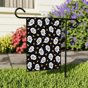 May include: A black garden flag featuring a repeating pattern of white cartoon ghosts and orange, yellow, and white candy corn. Each ghost has the text "TRUMP SUCKS" printed on its body. The flag is suspended from a black metal stand.