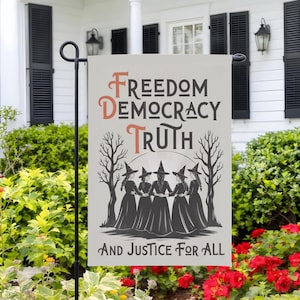 May include: A garden flag with the words "Freedom, Democracy, Truth, And Justice For All" in a stylized font. The flag features a graphic of five witches in black dresses and hats, standing in a circle, with bare trees on either side.