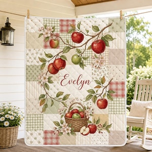 May include: A quilted blanket featuring a patchwork design of cream, green, and red plaid squares. The center displays a printed illustration of apple branches with red apples, green leaves, and white blossoms, with the name Evelyn in script.
