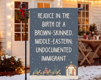 Può includere: Una bandiera da giardino blu scuro con il testo "REJOICE IN THE BIRTH OF A BROWN-SKINNED, MIDDLE-EASTERN; UNDOCUMENTED IMMIGRANT." La bandiera presenta un'illustrazione della scena della natività e tre cammelli.