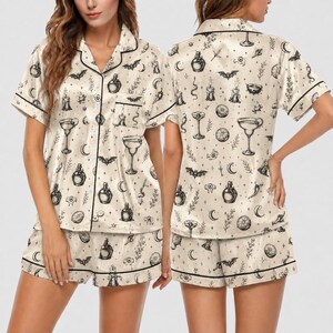 May include: A two-piece pajama set in cream satin featuring a black line-art print of bats, candles, potion bottles, crescent moons, and cocktail glasses. The short-sleeved button-down shirt has a notched collar, and the shorts have a matching trim.