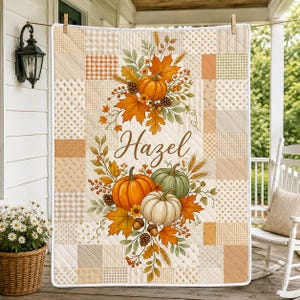 May include: A quilted blanket featuring a patchwork pattern in shades of beige, cream, and orange. The center displays a printed design of autumn leaves, wheat, pinecones, and three pumpkins with the name Hazel written in brown script text.