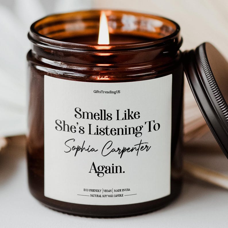 May include: A lit brown glass jar candle with a black lid. The candle has a white label with the text "Smells Like She's Listening To Sophia Carpenter Again." The candle is made of natural soy wax.