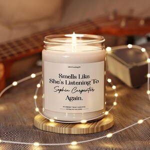 May include: A lit candle in a clear glass jar with the text "Smells Like She's Listening To Sophia Carpenter Again." The candle is on a wooden coaster, with fairy lights around it. A guitar is visible in the background.