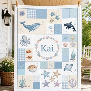 May include: A quilted blanket featuring a patchwork design with light blue and cream squares. The quilt displays illustrations of sea creatures including a whale, orca, octopus, pufferfish, stingray, and sea turtles, with the name Kai in the center.