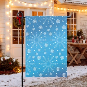 May include: A blue garden flag with a white snowflake pattern. The flag is displayed outdoors on a black metal stand. The background shows a house with a wreath and Christmas decorations.