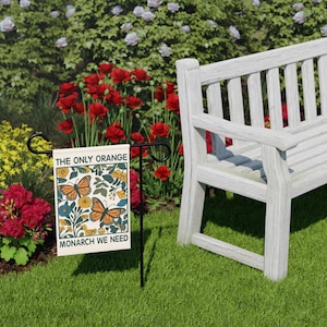 May include: A garden flag with the text "THE ONLY ORANGE MONARCH WE NEED" and a butterfly design. The flag is on a black metal stand, set in a grassy area with a white bench and red flowers in the background.