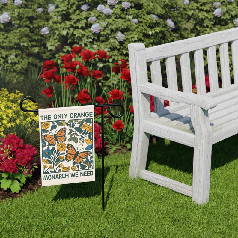 May include: A garden flag with the text "THE ONLY ORANGE MONARCH WE NEED" and a butterfly design. The flag is on a black metal stand, set in a grassy area with a white bench and red flowers in the background.