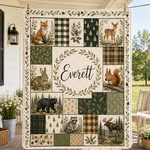 May include: A rectangular quilt featuring a central wreath surrounding the name Everett in black script. The quilt is composed of various panels displaying a fox, deer, rabbit, squirrel, bear, raccoon, and moose, alongside pine trees and plaid patterns.