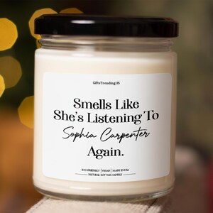 May include: A white candle in a glass jar with a black lid. The label reads "Smells Like She's Listening To Sophia Carpenter Again." The candle is eco-friendly, vegan, and made in the USA. It is a natural soy wax candle.