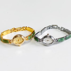May include: Two oval-faced quartz wristwatches with link bracelets. One watch is gold-toned with a gold dial, and the other is silver-toned with a white dial. Both bracelets feature alternating polished metal links and smooth, rectangular green stone inlays.