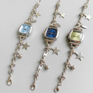 Peut inclure: Trois montres-bracelets argentées avec des cadrans carrés et des cadrans colorés : bleu clair, bleu foncé et vert clair. Chaque montre a une chaîne délicate avec des breloques en forme d'étoile et de boule. Les montres sont présentées sur un fond gris clair.