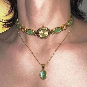 Op de afbeelding: Een goudkleurig choker-horloge met een ovale wijzerplaat en schakels van lichtgroene cabochonstenen, gecombineerd met een bijpassende goudkleurige ketting met een enkele ovale hanger van een lichtgroene cabochonsteen in het midden.