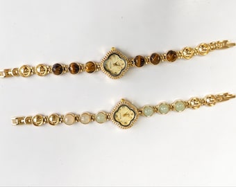 Op de afbeelding: Twee goudkleurige polshorloges met klavervormige kasten en schakelarmbanden. Het bovenste horloge heeft accenten van bruine tijgeroogsteen, terwijl het onderste horloge accenten van lichtgroene stenen heeft. Beide wijzerplaten hebben gouden wijzers.