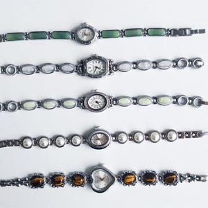 May include: Five silver-tone wristwatches arranged vertically. The top watch has green rectangular links. The second has oval white links. The third has pale green oval links. The fourth has white pearl-like links. The bottom watch has a heart-shaped face and brown tiger-eye stone links.