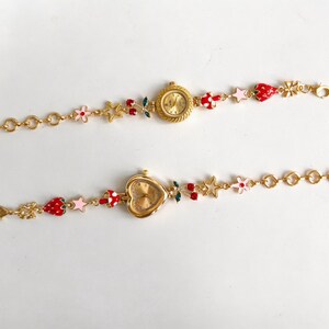 Peut inclure: Deux montres-bracelets dorées avec des breloques décoratives. L'une a un cadran rond, l'autre en forme de cœur. Les breloques comprennent des fraises, des fleurs, des étoiles et des cerises. Les bracelets sont en maillons dorés.