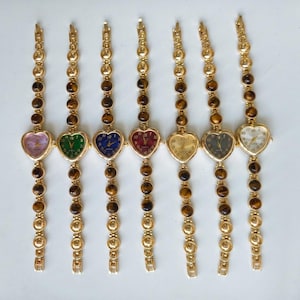 May include: Seven gold-tone wristwatches arranged in a row. Each watch features a heart-shaped face with a unique dial color including purple, green, blue, red, gold, black, and white. The bands consist of round tiger eye stone beads.