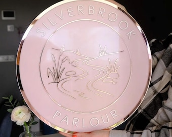 Puede incluir: Un letrero circular con fondo rosa pálido y borde de oro rosa metálico. El texto "SILVERBROOK" está arqueado en la parte superior y "PARLOUR" en la inferior en letras en relieve. Un dibujo lineal central representa un arroyo sinuoso.