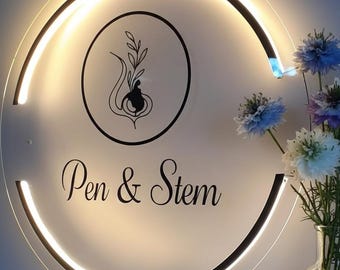 Puede incluir: Un letrero circular iluminado con el texto "Pen & Stem" en escritura elegante. El letrero presenta un gráfico en blanco y negro dentro de un círculo, y está rodeado por una luz brillante. Un jarrón de flores azules y blancas está a la derecha.