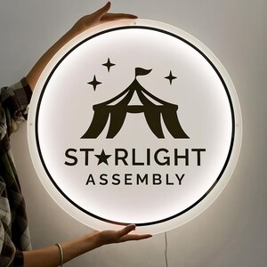 May include: A round, illuminated sign with a white background and a black border. The sign features a black circus tent graphic with the words "STARLIGHT ASSEMBLY" in black text. Three black stars are above the tent graphic.