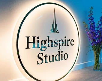 May include: A round, illuminated sign with the text "Highspire Studio" in black lettering. The sign has a white face with a black border and a teal logo. A power cord extends from the bottom.
