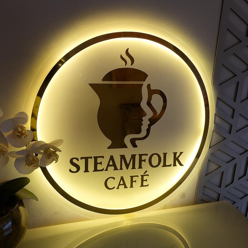 May include: A round, illuminated sign with a gold border and a cream-colored center. The sign features a gold silhouette of a teapot with a face profile and the words "STEAMFOLK CAF&Eacute;" in dark letters. The sign is lit with a warm, inviting glow.