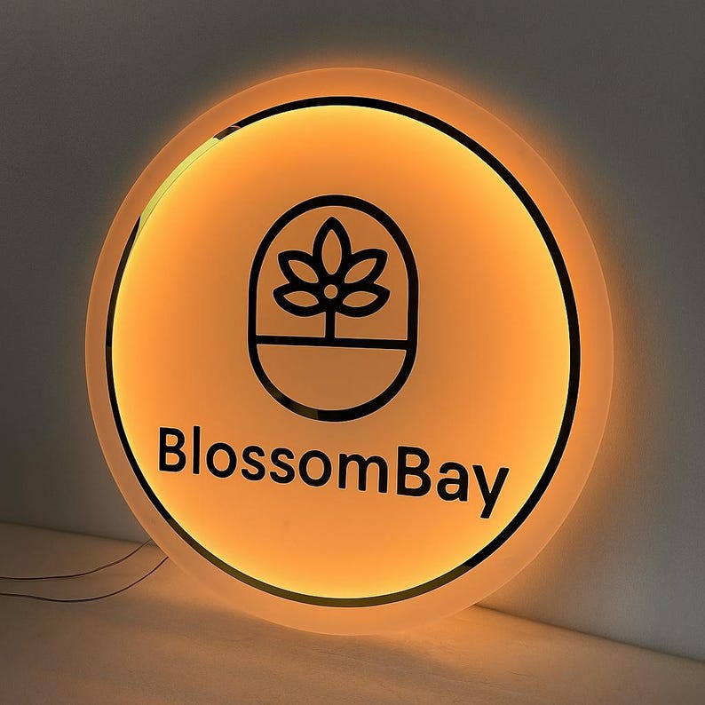 May include: A round, illuminated sign with a warm orange glow. The sign features a black outline of a flower and the text "BlossomBay" in a modern font. The sign is likely for a business.