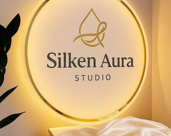 Puede incluir: Un letrero circular iluminado con un borde dorado y las palabras "Silken Aura STUDIO" en una fuente serif. El letrero tiene un emblema dorado en la parte superior. Una tela de satén blanca está drapeada debajo.