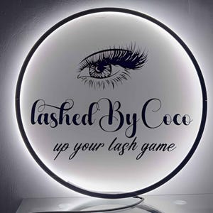 May include: A round, illuminated sign with a black border. The sign features a stylized eye with long lashes and the text "Lashed By Coco" and "up your lash game" in a script font. The background is white.