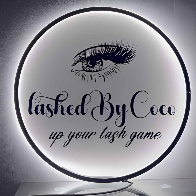 May include: A round, illuminated sign with a black border. The sign features a stylized eye with long lashes and the text "Lashed By Coco" and "up your lash game" in a script font. The background is white.