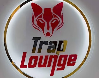 Puede incluir: Un letrero redondo e iluminado con un logotipo de cabeza de zorro rojo sobre las palabras "Trap Lounge" en letras rojas y negras. El letrero tiene un borde dorado y un fondo blanco, creando un contraste llamativo.