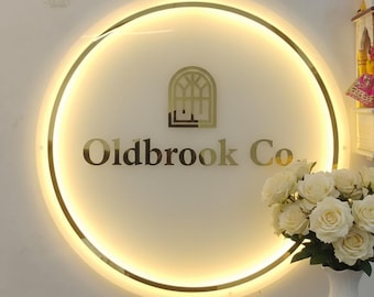 Puede incluir: Un letrero redondo e iluminado con un borde dorado y el texto "Oldbrook Co." en letras doradas. El letrero presenta un diseño arquitectónico dorado encima del texto. Un jarrón de rosas blancas se encuentra debajo.
