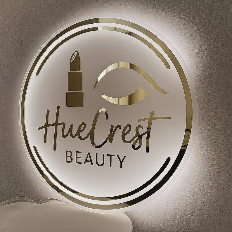 May include: A round, illuminated sign with a gold and white design. The sign features a lipstick, an eye, and the words "HueCrest BEAUTY" in gold lettering. The sign is surrounded by a gold border and a white light.