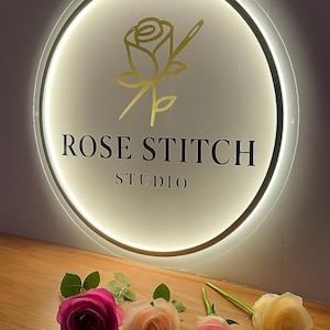 Może przedstawiać: Okrągły, podświetlany szyld z napisem "ROSE STITCH STUDIO" i złotym logo r&oacute;ży. Poniżej, cztery r&oacute;że w odcieniach r&oacute;żu i ż&oacute;łci są ułożone na drewnianej powierzchni.