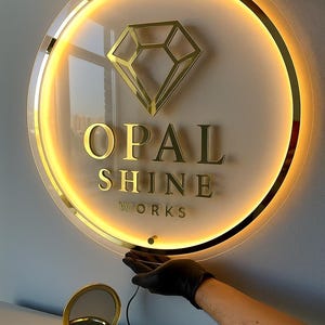Może przedstawiać: Okrągły, podświetlany szyld ze złotym logo diamentu i napisem "OPAL SHINE WORKS". Znak ma złotą obw&oacute;dkę i ciepłe, świecące światło. Małe, złote lusterko kompaktowe znajduje się na pierwszym planie.