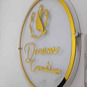 May include: A round, clear acrylic sign with a gold border and gold lettering. The sign says "Dream Cosmetics" and features a gold silhouette of a woman's face with flowers.