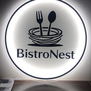 Peut inclure: Enseigne circulaire BistroNest avec un fond blanc &eacute;clair&eacute;. L'enseigne pr&eacute;sente un graphisme noir d'une fourchette et d'une cuill&egrave;re au-dessus d'un nid stylis&eacute;, avec les mots "BistroNest" en gras, en police sans-serif.