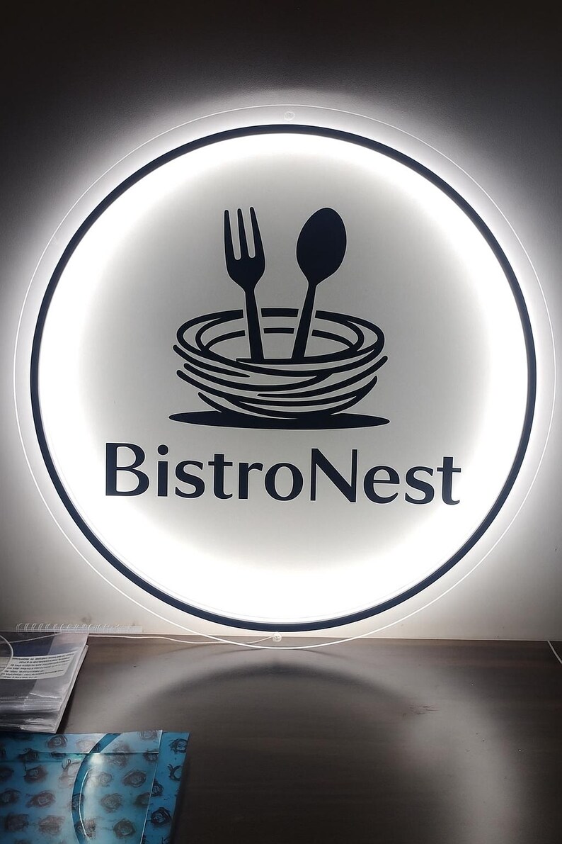 Peut inclure: Enseigne circulaire BistroNest avec un fond blanc &eacute;clair&eacute;. L'enseigne pr&eacute;sente un graphisme noir d'une fourchette et d'une cuill&egrave;re au-dessus d'un nid stylis&eacute;, avec les mots "BistroNest" en gras, en police sans-serif.