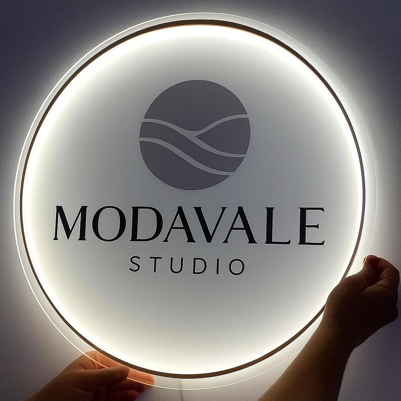 May include: A round, illuminated sign with the text "MODAVALE STUDIO" in black. The sign has a white face with a gray logo and a bright, white light around the edge. The sign is being held by a person.