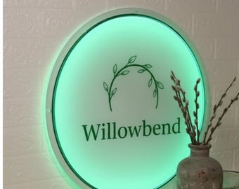 Puede incluir: Un letrero redondo e iluminado con la palabra "Willowbend" en letras verdes. El letrero tiene un brillo verde y un diseño de hojas decorativas. Un jarrón con ramas está en primer plano.