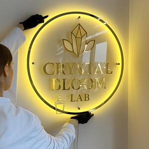 Może przedstawiać: Okrągły, podświetlany szyld z napisem "CRYSTAL BLOOM LAB" w złotych literach. Znak posiada złotą obw&oacute;dkę oraz wz&oacute;r kryształu i liści. Tło jest ciepło, jasnoż&oacute;łte.