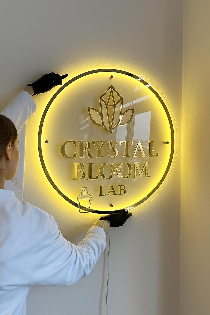 Może przedstawiać: Okrągły, podświetlany szyld z napisem "CRYSTAL BLOOM LAB" w złotych literach. Znak posiada złotą obw&oacute;dkę oraz wz&oacute;r kryształu i liści. Tło jest ciepło, jasnoż&oacute;łte.