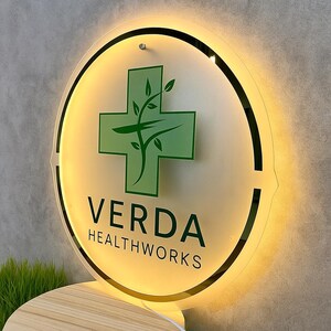 May include: A round, illuminated sign with the text "VERDA HEALTHWORKS". The sign features a green cross with a plant design in the center. The sign has a black border and is mounted on a wall.