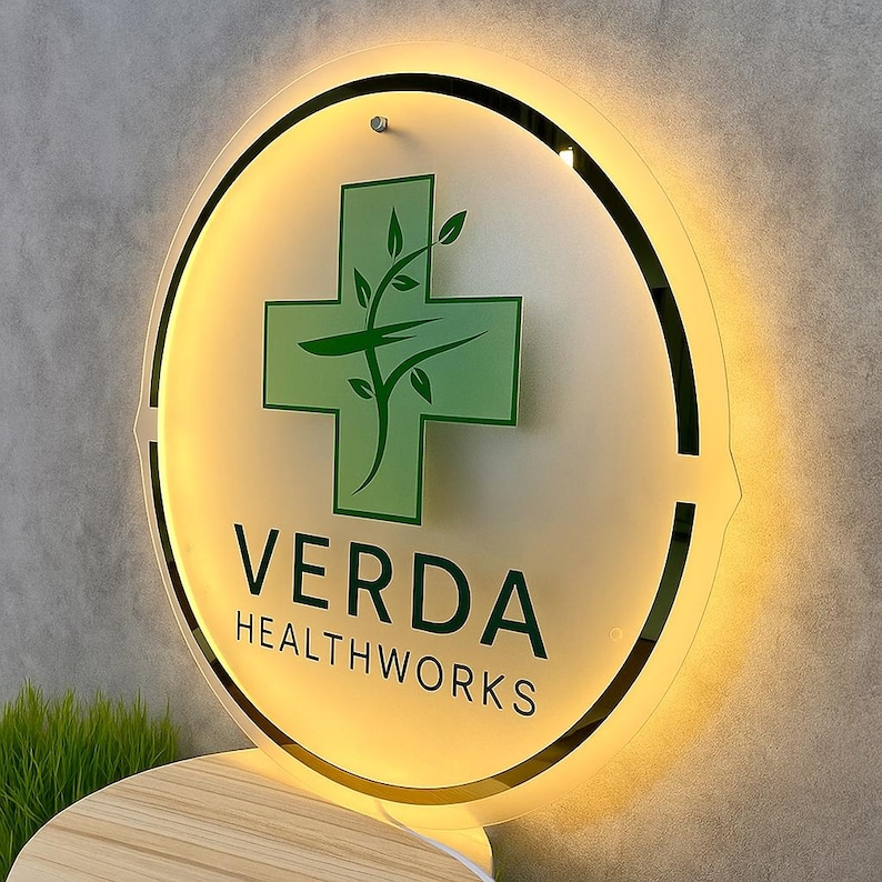 May include: A round, illuminated sign with the text "VERDA HEALTHWORKS". The sign features a green cross with a plant design in the center. The sign has a black border and is mounted on a wall.