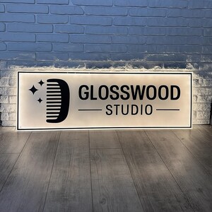 May include: Rectangular illuminated sign with the text "GLOSSWOOD STUDIO" in black. The sign features a comb graphic and star icons on the left. The sign is mounted on a white brick wall above a wooden floor.