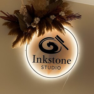 May include: A round, illuminated sign with the text "Inkstone STUDIO" in black. The sign features a black graphic and is surrounded by a warm, glowing light. A decorative floral arrangement of dried plants and grasses is above the sign.